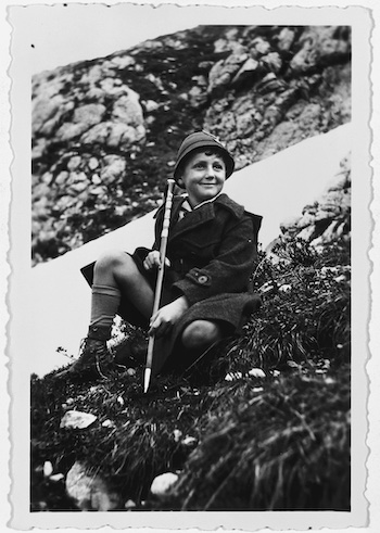 Young boy sitting on a hill wearing a hat and coat and wearing a stick.