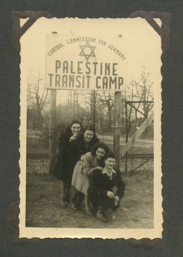 a black and white photograph featuring four smiling people, with a sign behind them that reads "Control Commission for Germany, Palestine Transit Camp"