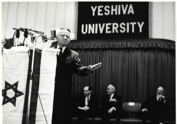 black and white photo of white man behind a podium with an Israeli flag, behind him men on chairs and a sign saying "Yeshiva University"