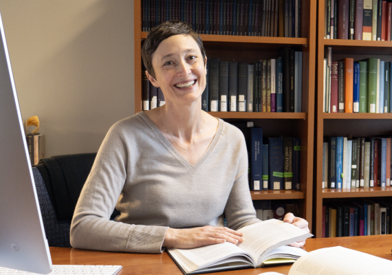Short-haired white woman smiles while sitting at a desk with a book.