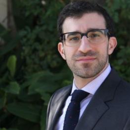 Man with horn-rimmed glasses and stubble looks over his left shoulder at the viewer.