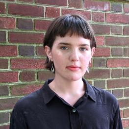 Photo of a person with short hair in a black button down shirt in front of a brick wall