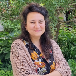 Smiling woman with long hair stands in a garden with her arms folded.