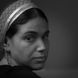 Woman wearing headscarf and dangling earrings looks towards the viewer from over her right shoulder.