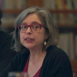 Woman with bobbed hair and glasses looks to her left opening her mouth, about to speak.