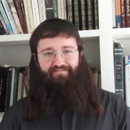 A bearded man with glasses in front of a bookcase.