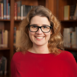 A woman with wavy hair and rectangular glasses smiles as she stands in front of a bookcase.