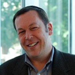 Photograph of man tilting his head and smiling, wearing a suit jacket and an open-collared shirt.